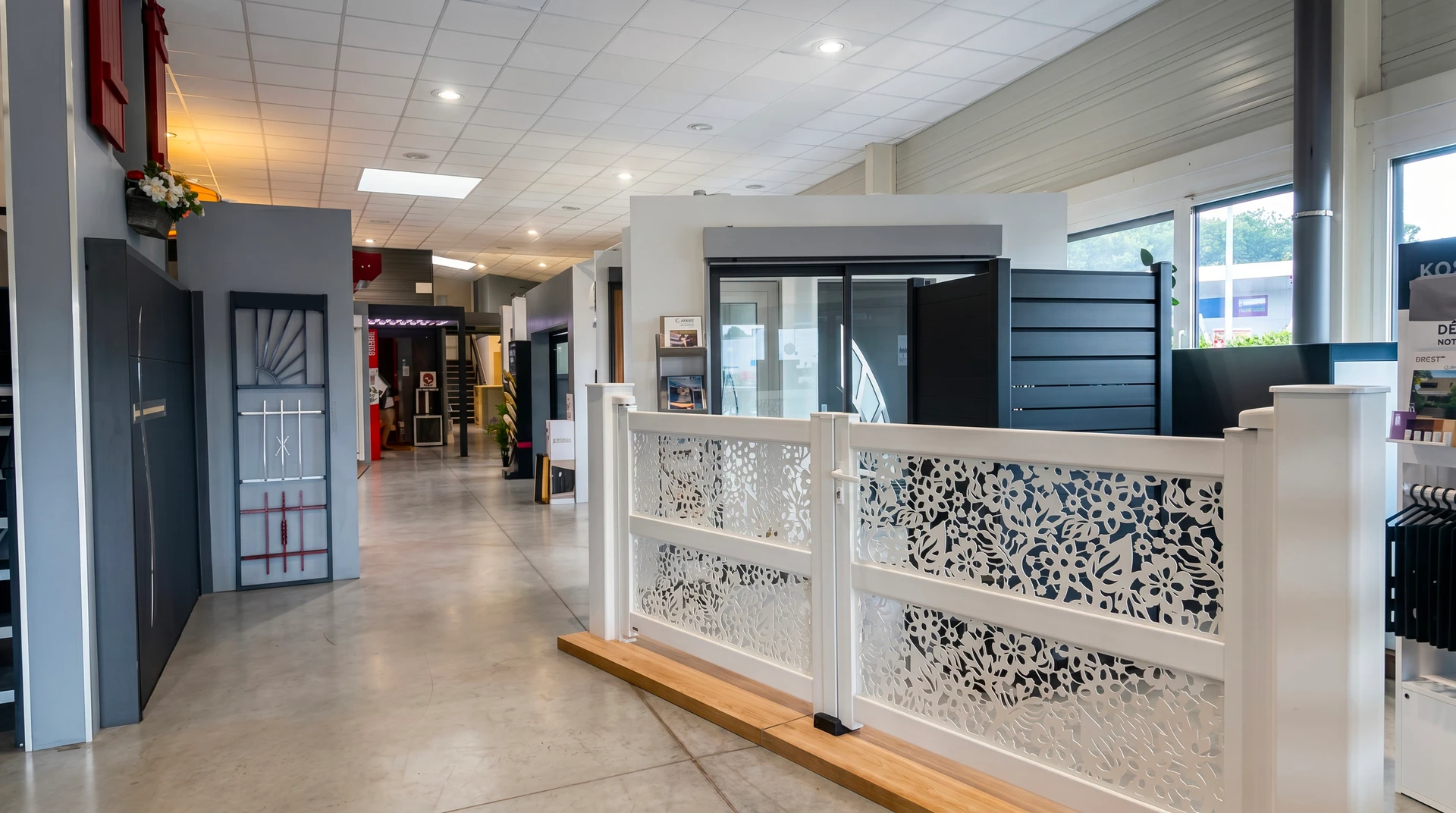 Photographie du showroom de Tino' Trans Fermetures montrant un espace spacieux et modernisé avec une pergola bioclimatique et un carport présentés grandeur nature côte à côte. En avant-plan, une borne interactive ou un bureau de consultation avec un écran tactile permettant la personnalisation des produits. L'espace est lumineux, bien aménagé, avec des vitrines exposant des fenêtres, portes et portails en différents matériaux (PVC, aluminium, bois). Un ou deux clients ou vendeurs peuvent être visibles en arrière-plan interagissant naturellement dans ce contexte professionnel.