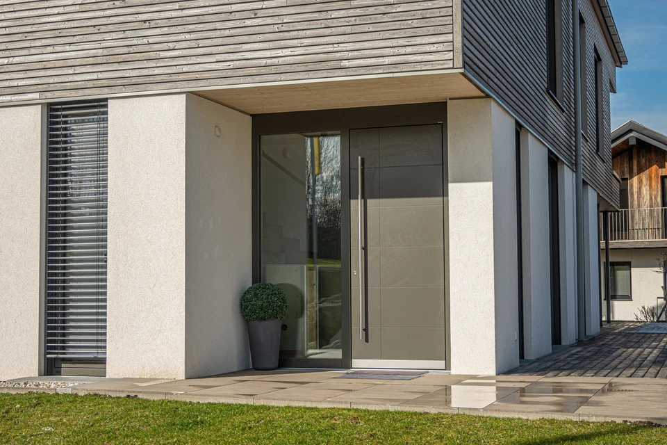Porte d'entrée en aluminium moderne de haute qualité, photographiée de face depuis l'extérieur d'une maison résidentielle contemporaine. La porte présente un design épuré et élégant avec des vitrages ou panneaux modernes, intégrée harmonieusement à la façade du bâtiment. L'image doit transmettre à la fois la robustesse (protection), l'isolation performante et l'esthétique soignée. La mise en lumière naturelle souligne la qualité du produit et son intégration dans un environnement résidentiel prestigieux. Aucun technicien, aucun travail en cours, uniquement le produit fini dans son contexte d'utilisation.
