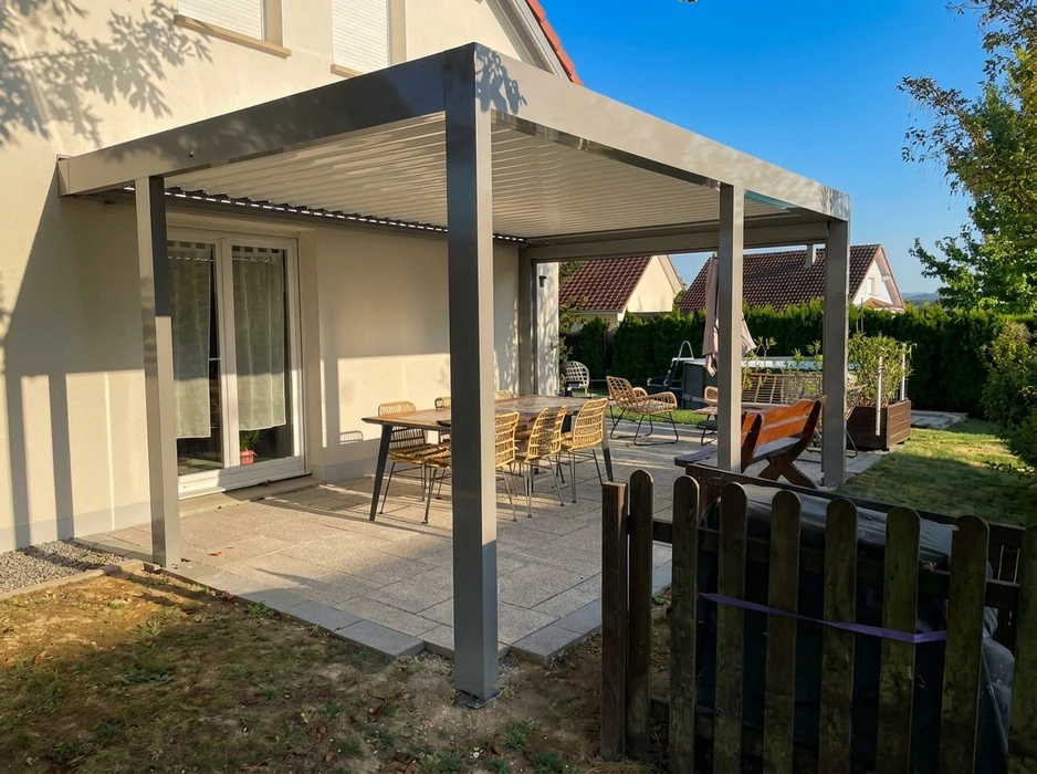 Terrasse moderne en dallage béton avec pergola bioclimatique aluminium anthracite à lames orientables, adossée à une maison contemporaine