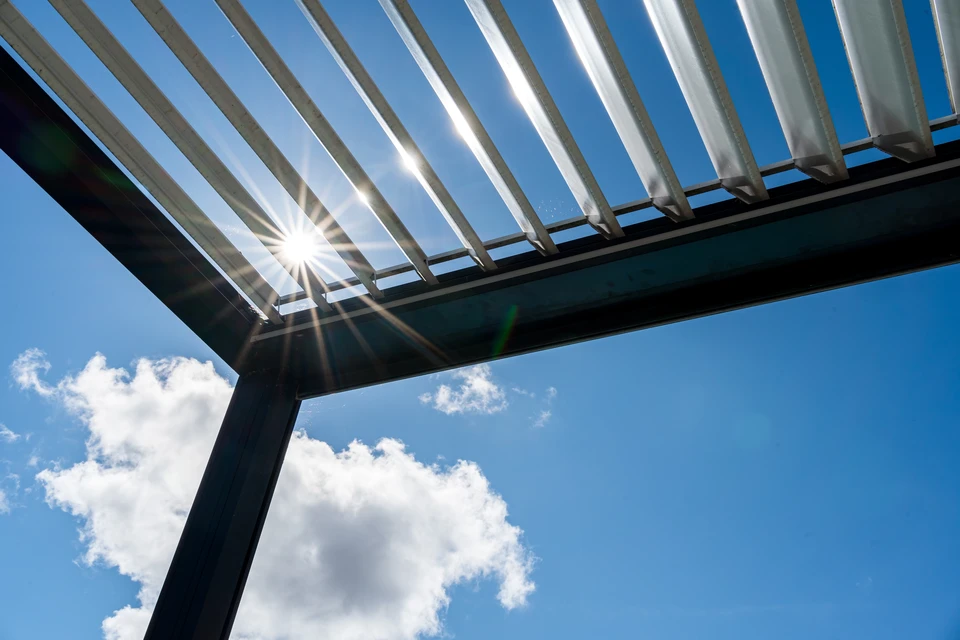 Photographie d'un showroom d'exposition extérieur ou semi-couvert présentant une pergola bioclimatique moderne avec lames orientables et un carport adjacent, tous deux en grandeur nature. Les structures sont visibles dans un espace de démonstration professionnel bien éclairé, permettant de voir les détails de construction, les coloris et les finitions. L'image doit montrer des structures fonctionnelles et attractives, idéales pour que les clients puissent visualiser et actionner les éléments mobiles.